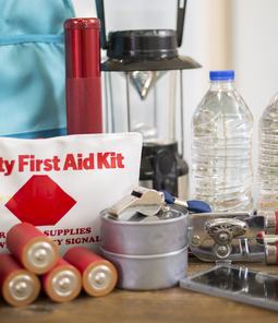 An emergency preparedness kit with a first aid bag, water bottles, batteries, a lantern, can opener and backpack on a table.