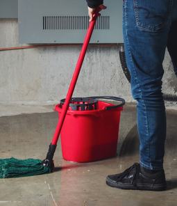 A person mopping standing water off a flooded basement floor with a red bucket nearby.